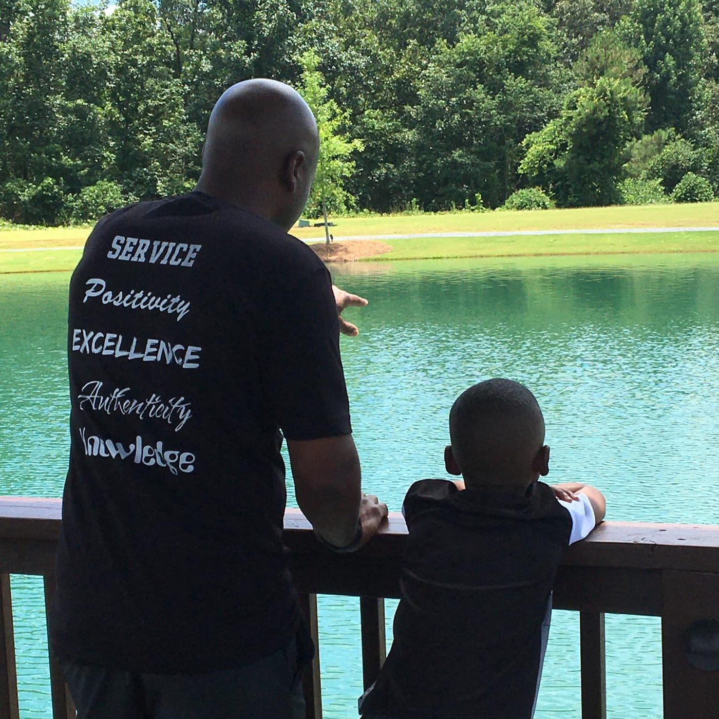 Neco Johnson and his son looking out over a lake, the back of Neco's shirt reading Service, Positivity, Excellence, Authenticity, Knowledge.